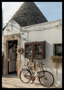 Plakat podróżniczy z białym budynkiem skąpanym w słońcu i stojącym obok rowerem - fotografia wykonana w popularnym Alberobello